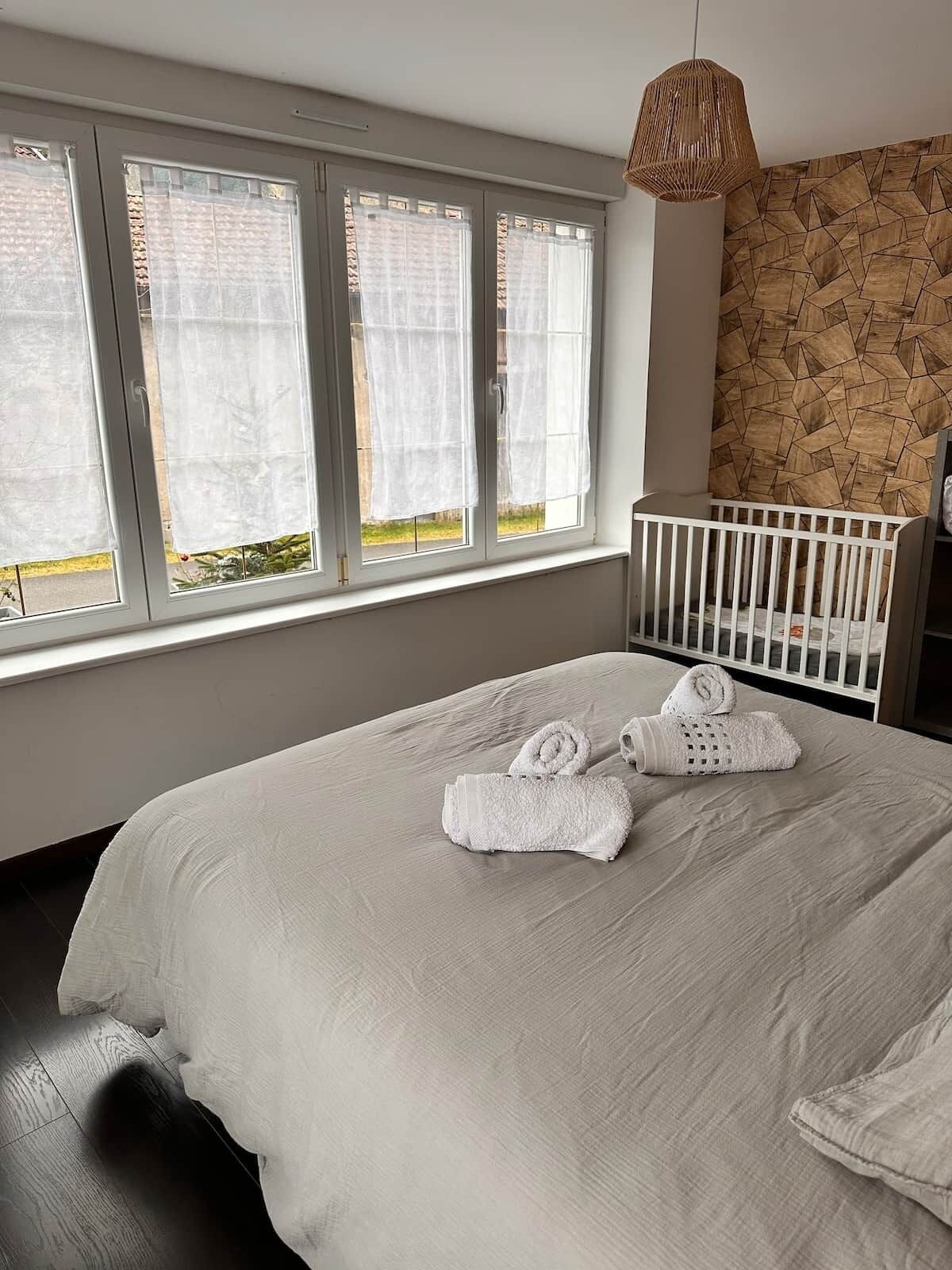 A bedroom with a large bed, white curtains, a crib, and a geometric-patterned wall.