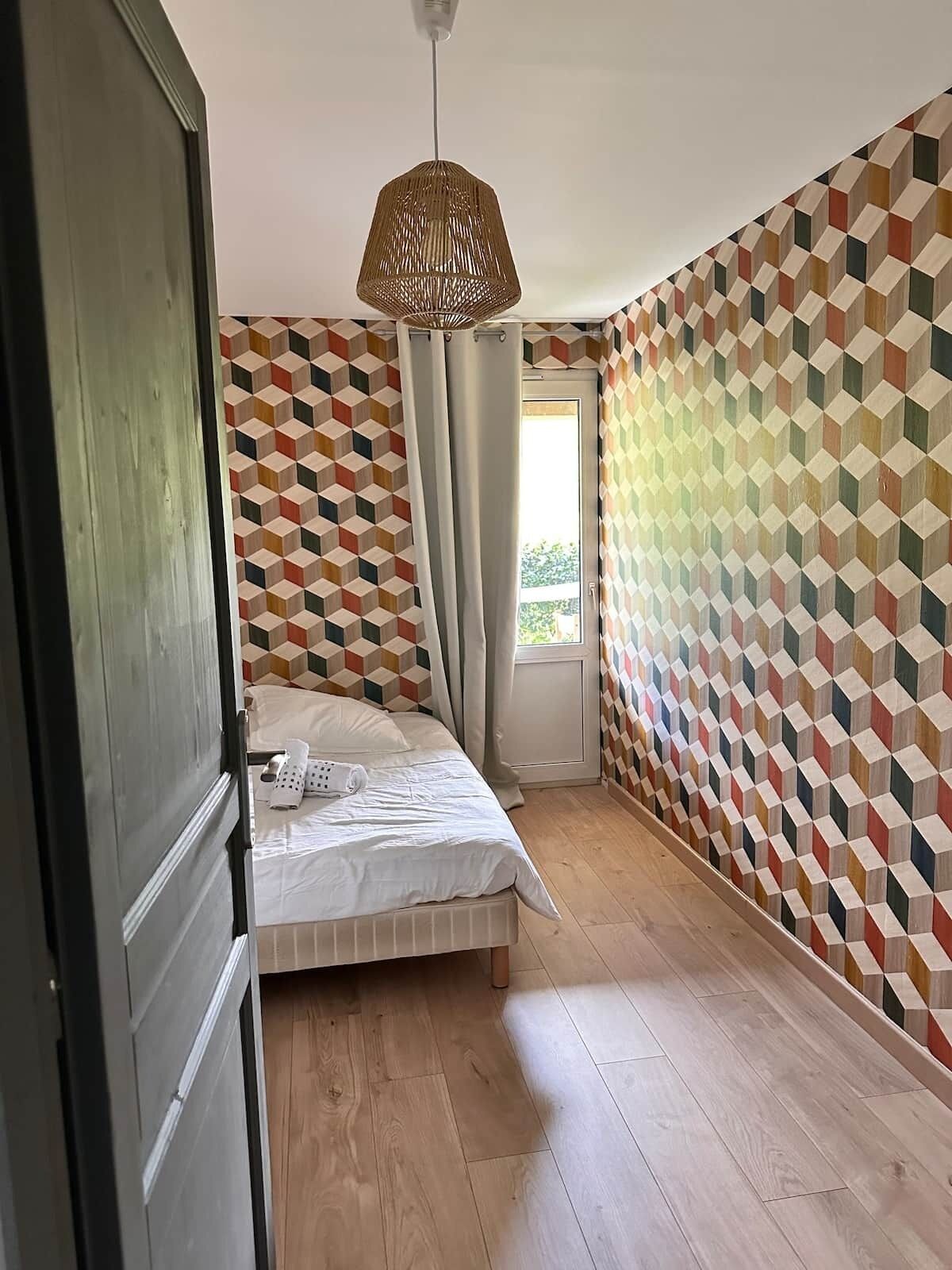 A bedroom with a geometric-patterned wall, a bed with white bedding, a hanging light fixture, and a window with curtains.