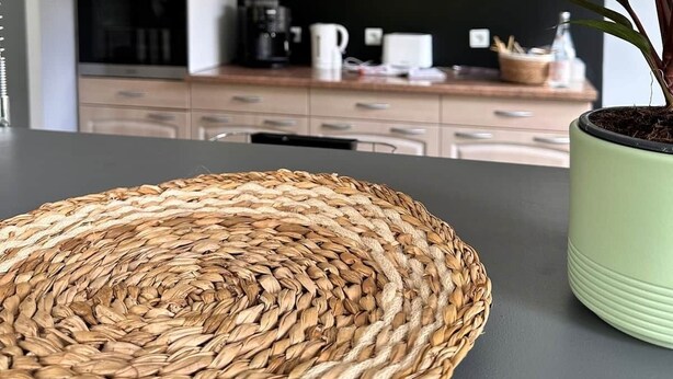 Une cuisine avec des meubles de couleur bois clair, un set de table en fibres tressées, une plante verte en pot et un luminaire suspendu.