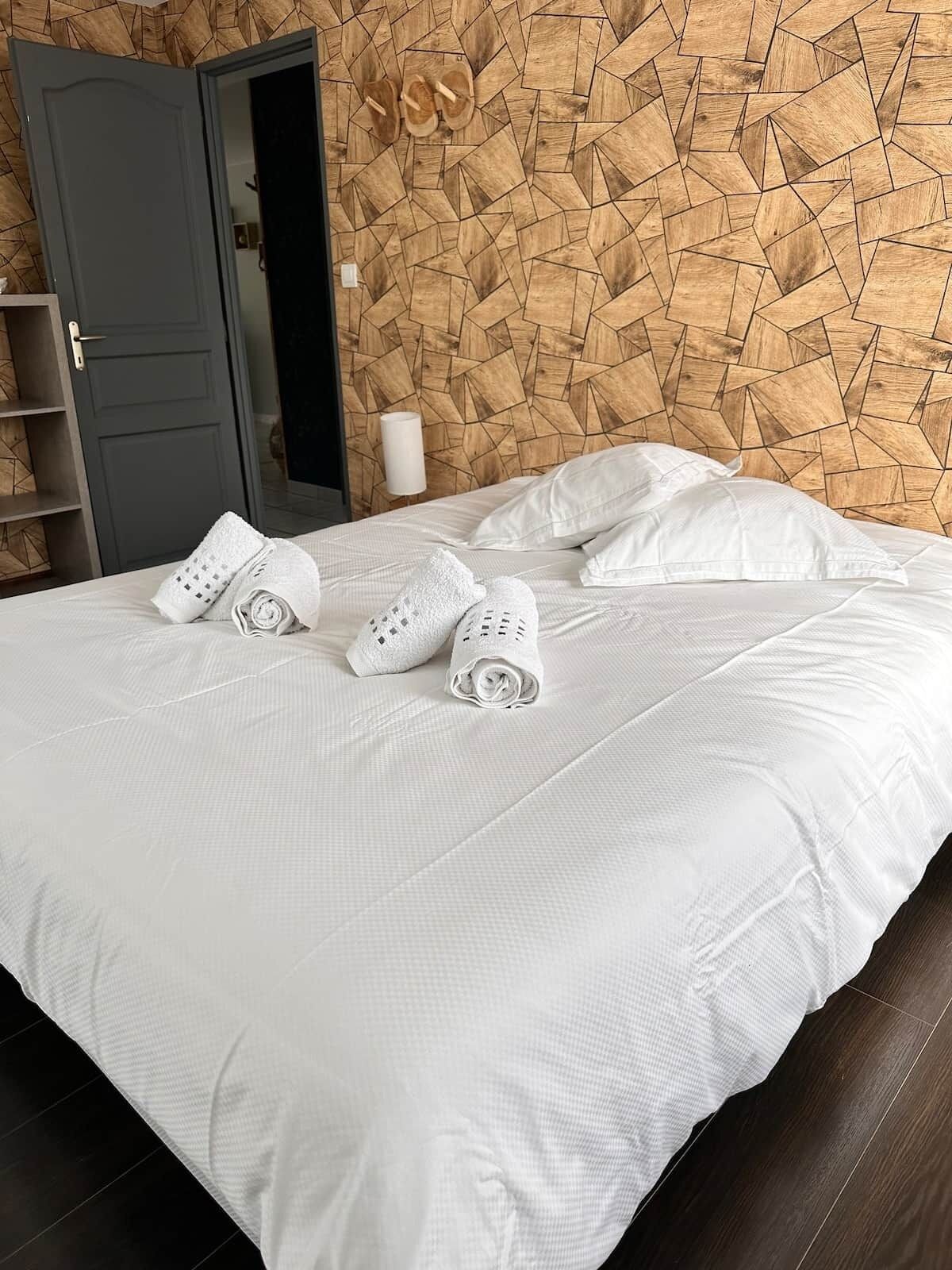 A neatly made bed with white linens and towels, a geometric wooden headboard, a dark door, and a white wall-mounted lamp.