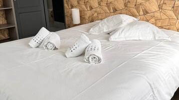 A neatly made bed with white linens and towels, a geometric wooden headboard, a dark door, and a white wall-mounted lamp.