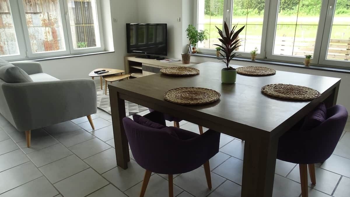 A modern living room with a dining table, purple chairs, a sofa, a TV, and large windows with a view of greenery.