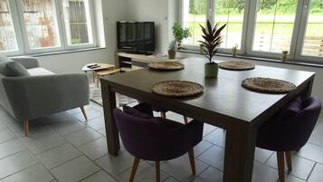 A modern living room with a dining table, purple chairs, a sofa, a TV, and large windows with a view of greenery.