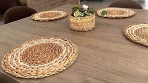 Un coin repas avec une table en bois, des sets de table en fibres tressées et un grand panier tressé placé au centre, contenant des plantes vertes.