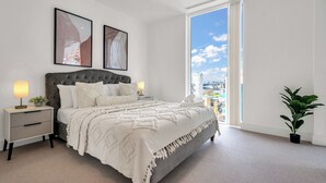 Een slaapkamer met een groot bed, een nachtkastje, een potplant en een stadszicht door een groot raam.