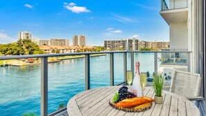 Un balcón con vista al río y al paisaje urbano, una mesa de madera con una botella de champán, uvas y una hogaza de pan.