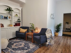 Ein gemütliches Wohnzimmer mit zwei Sesseln, einem Holztisch, einem gemusterten Teppich und einem Bücherregal voller Bücher und Dekoration.