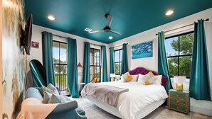 Une chambre à coucher comprenant un lit, une chaise, une table de chevet, un ventilateur de plafond et de grandes fenêtres avec des rideaux.