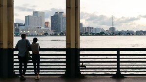 Un couple se tient sur une promenade en bord de mer, avec la silhouette de la ville en arrière-plan.