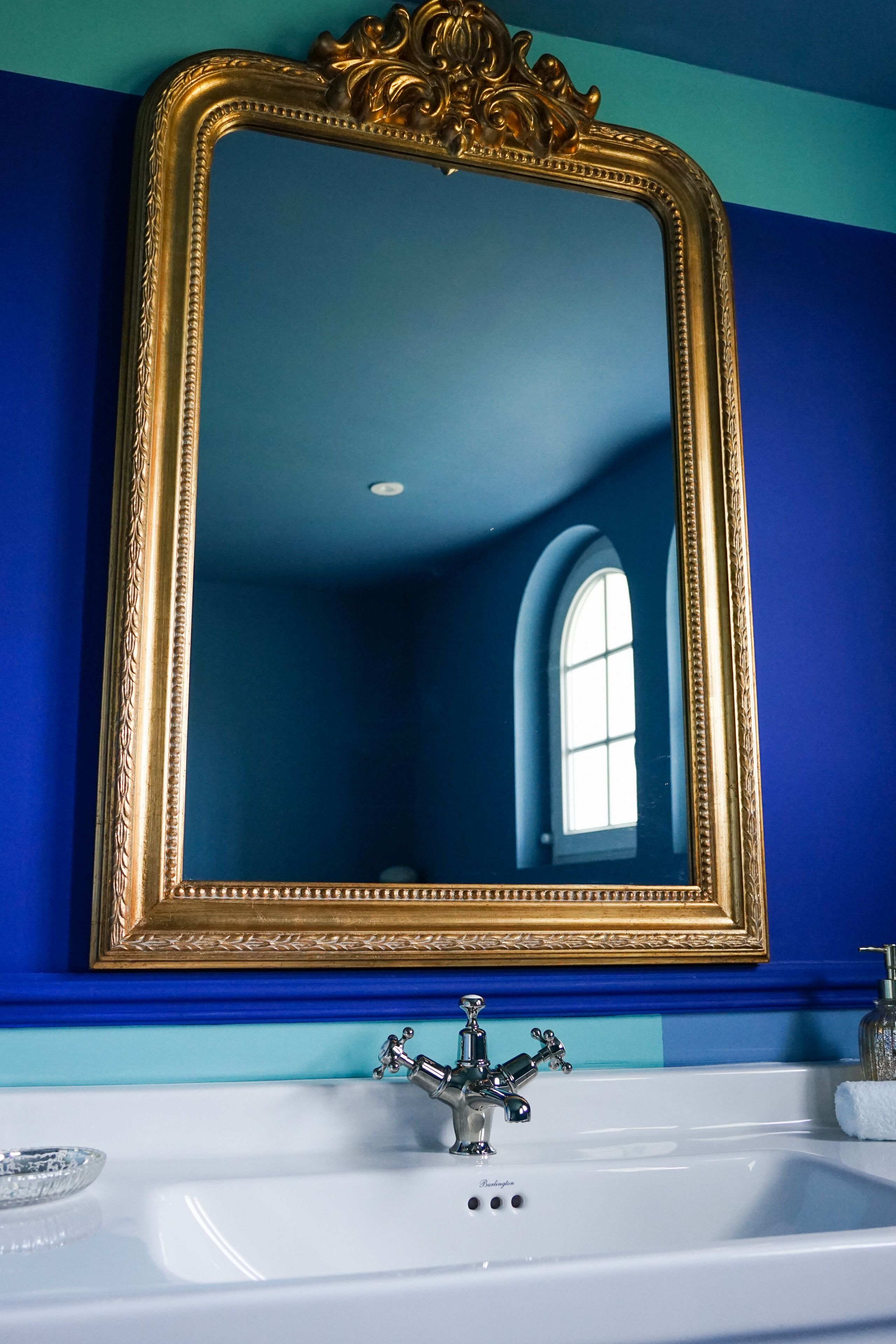 Un baño con un espejo de marco dorado, un lavamanos blanco y una pared azul.