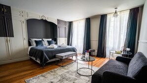 Ein modernes Schlafzimmer mit einem Doppelbett, einem grauen Sofa, einem kleinen Tisch mit Flaschen und einem großen Fenster mit Vorhängen.