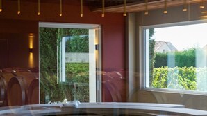Ein Raum mit einer runden Sitzecke, umgeben von Holzfässern, einem großen Fenster mit Blick ins Grüne und hängenden Pendelleuchten.