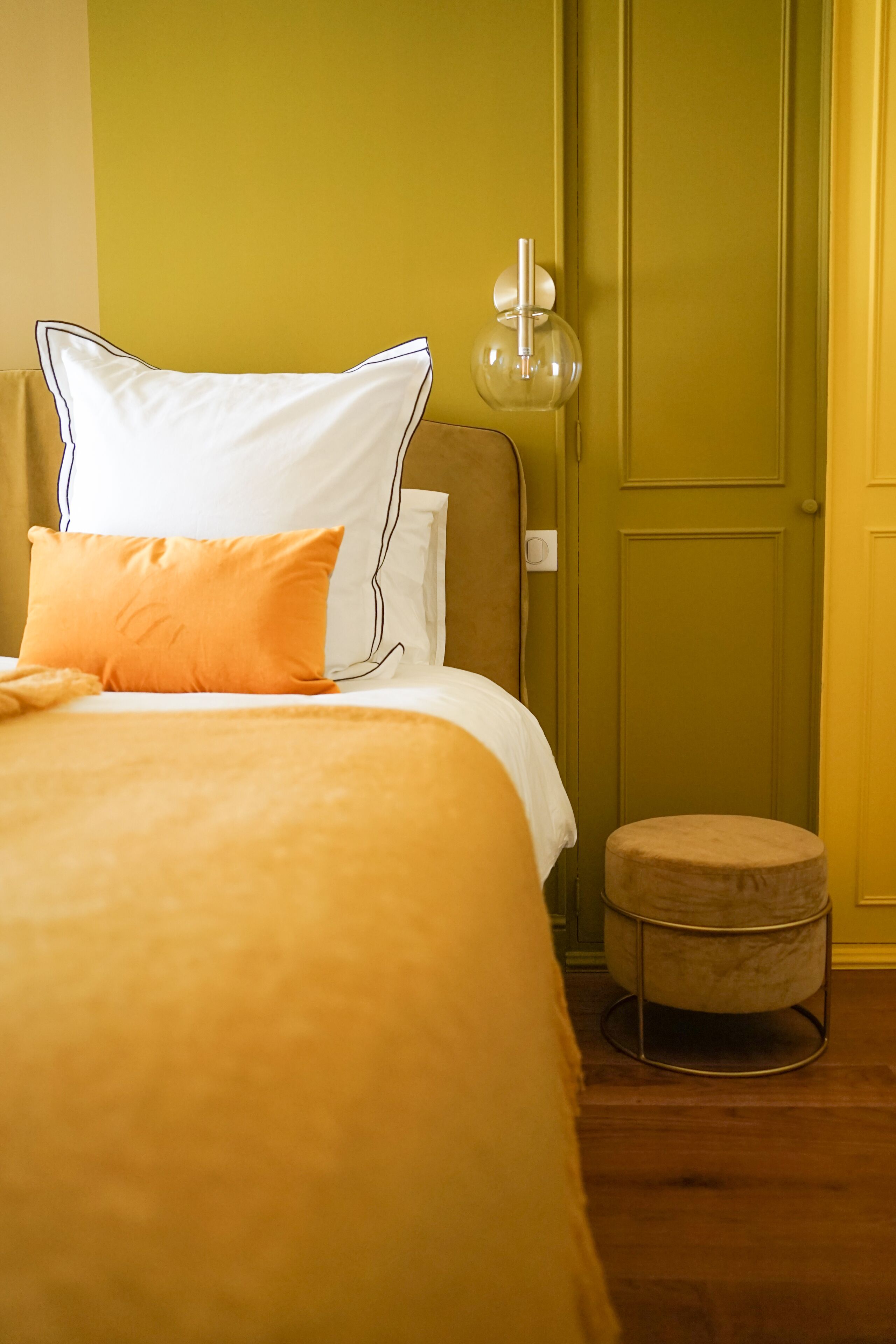 A bed with white and orange bedding, a round ottoman, a wall-mounted lamp, and a green door.