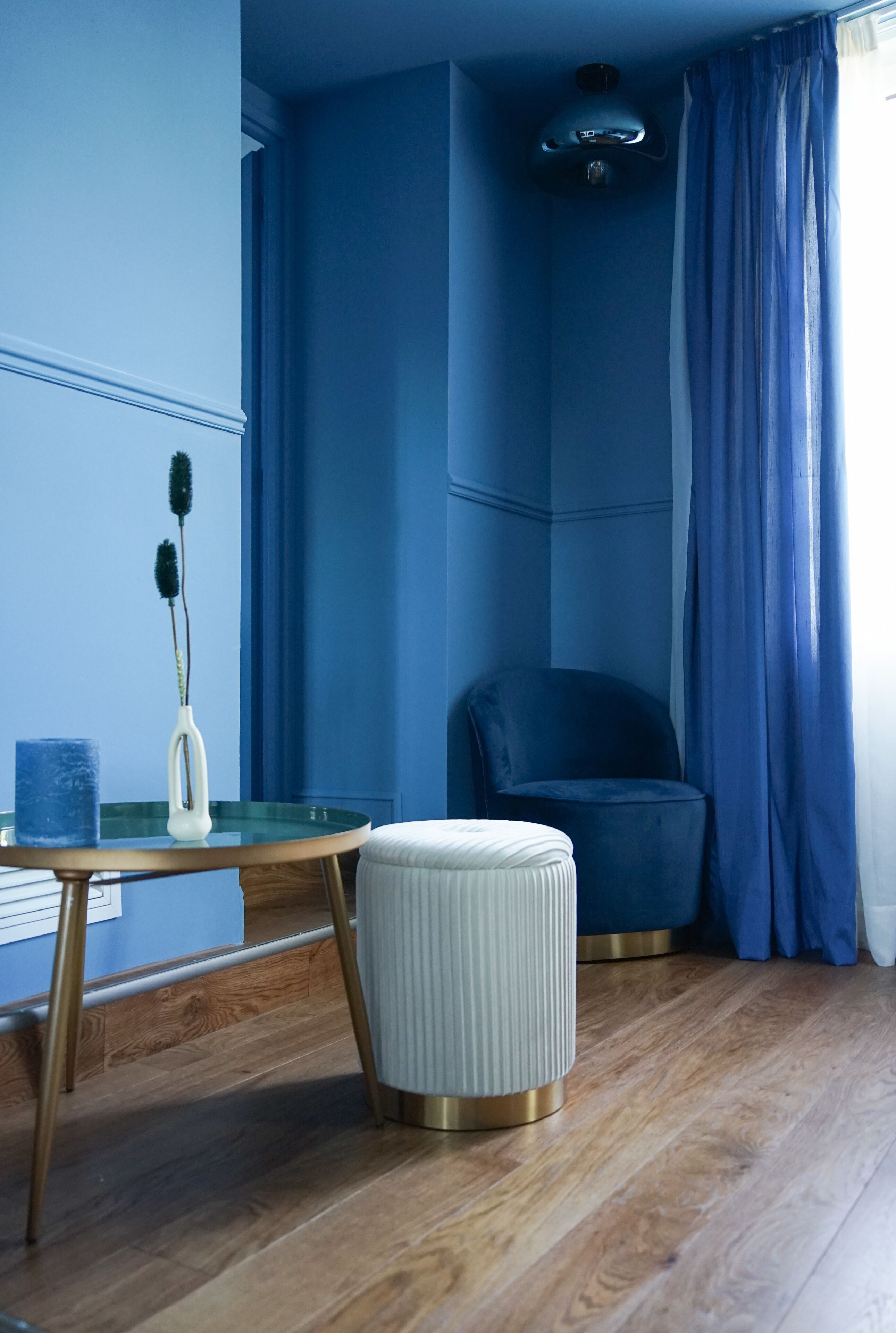 Ein Zimmer mit blauer Wand, einem runden Tisch mit einer Vase und einem blauen Behälter, einem weißen Hocker, einem blauen Sessel und einem Fenster mit blauen Vorhängen.