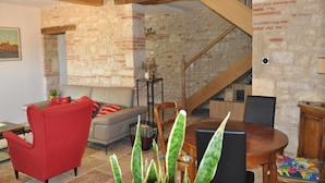 Un salon avec un fauteuil rouge, un canapé gris, une table à manger en bois avec des chaises, un escalier avec des rampes en bois et un mur en pierre.