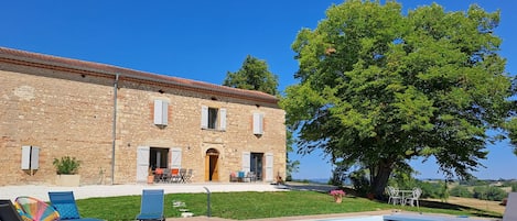 A stone building with a swimming pool, outdoor seating, and a large tree.