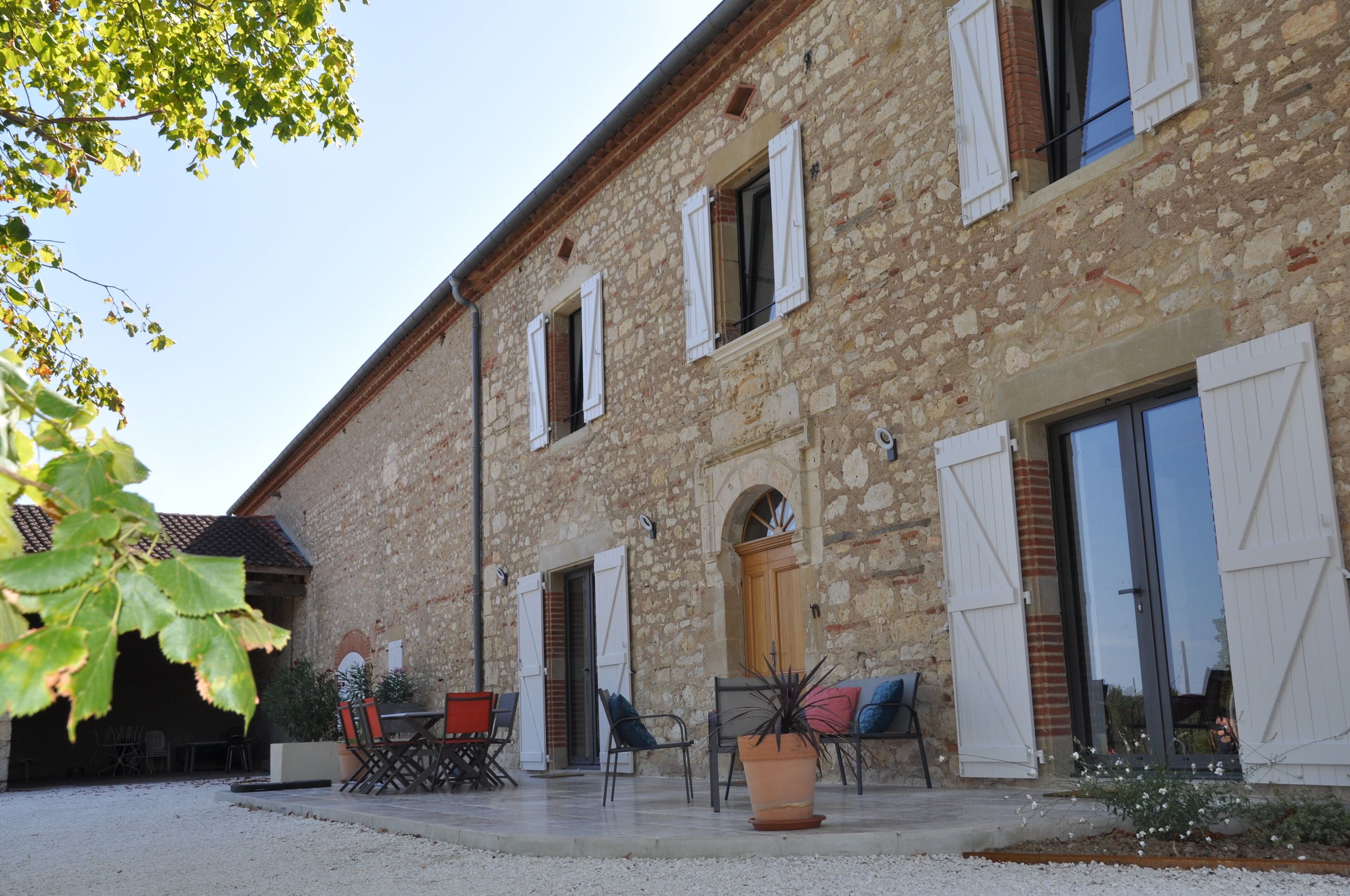 Un bâtiment en pierre avec des volets blancs, une porte en bois et un espace détente comprenant des chaises et une table.