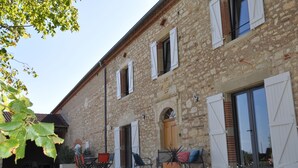 Un bâtiment en pierre avec des volets blancs, une porte en bois et un espace détente comprenant des chaises et une table.