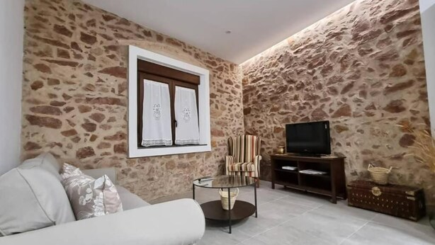 A living room with a stone wall, a sofa, a glass coffee table, a TV, and a wooden entertainment unit. - Tourist House in the center of Vilafamés (Vilafamés)