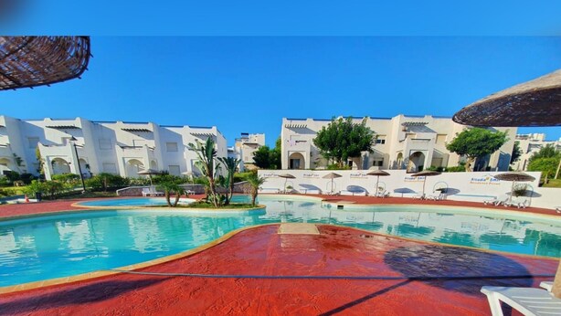A swimming pool with red flooring, white lounge chairs, and white buildings in the background. - La Vida Villa Alcudia Simr-Fnideq (Fnideq)