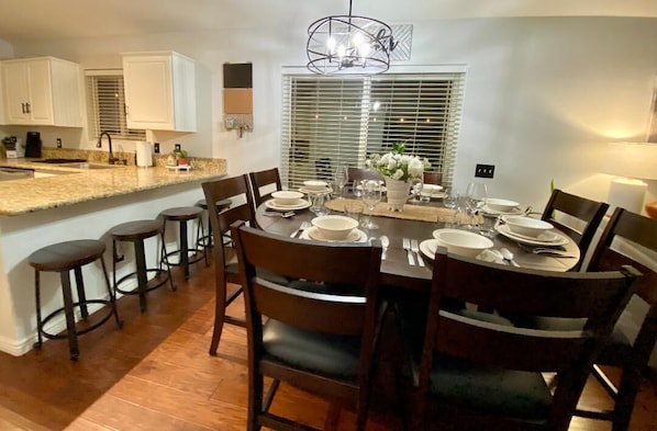 A dining area with a round table set for six, surrounded by wooden chairs. There is a kitchen area with white cabinets and a granite countertop.