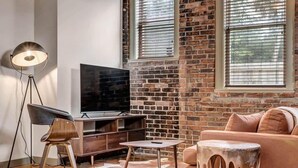 A modern living room with a brick wall, a flat-screen TV, a wooden entertainment unit, a leather sofa, a coffee table, and a floor lamp. - Modern-Industrial 2B Townhouse Close to DT! (Jacksonville)
