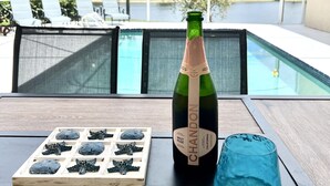 En flaske Brut Rose Chandon champagne, et blåt glas og en træbakke med dekorative genstande på et bord ved en pool med udsigt til palmer og en klar himmel.