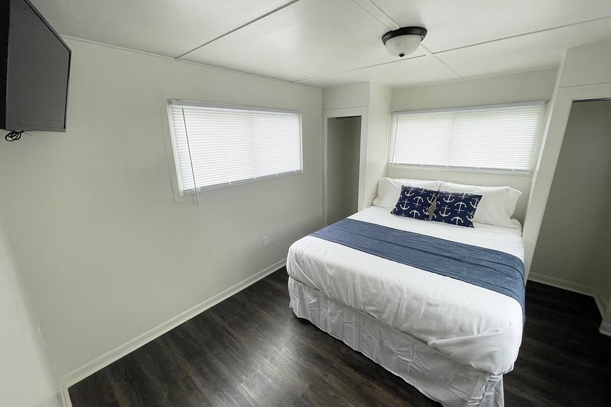 A bedroom with a bed, two windows, a ceiling fan, and a mounted TV.