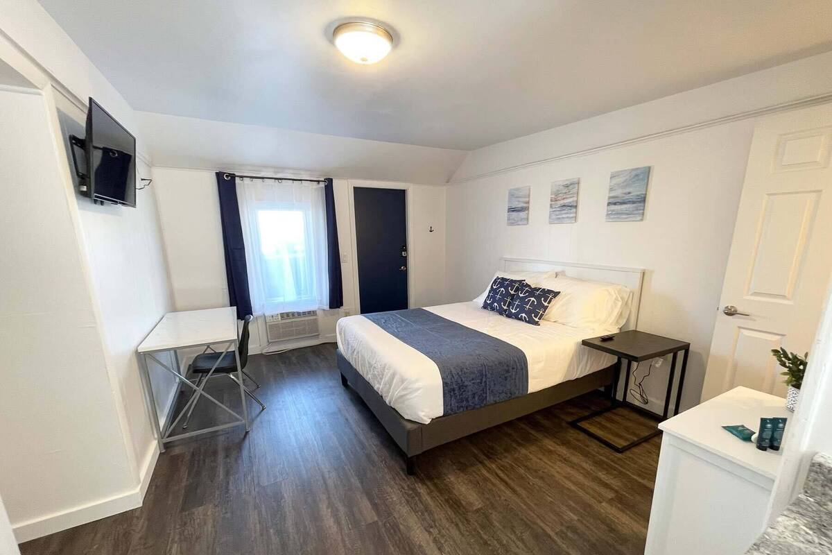 A bedroom with a bed, a desk, a chair, a small table, and a TV on the wall.