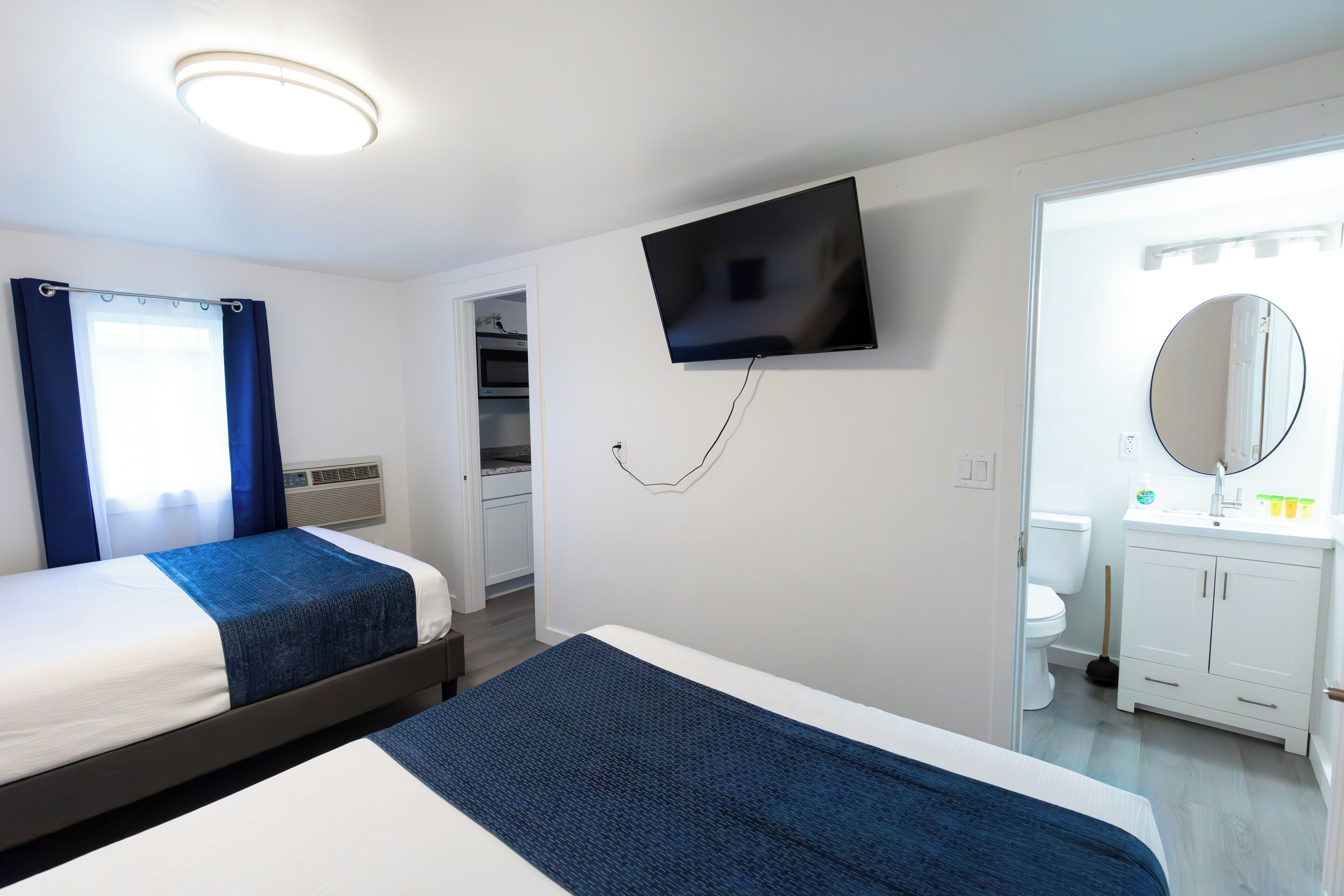 Habitación de hotel con dos camas, televisor, baño con espejo y vistas a una cocina y un balcón.