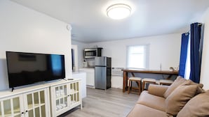 A modern living room with a flat-screen TV, a white cabinet with glass doors, a beige sofa, and a kitchen area with a microwave, refrigerator, and dining table.
