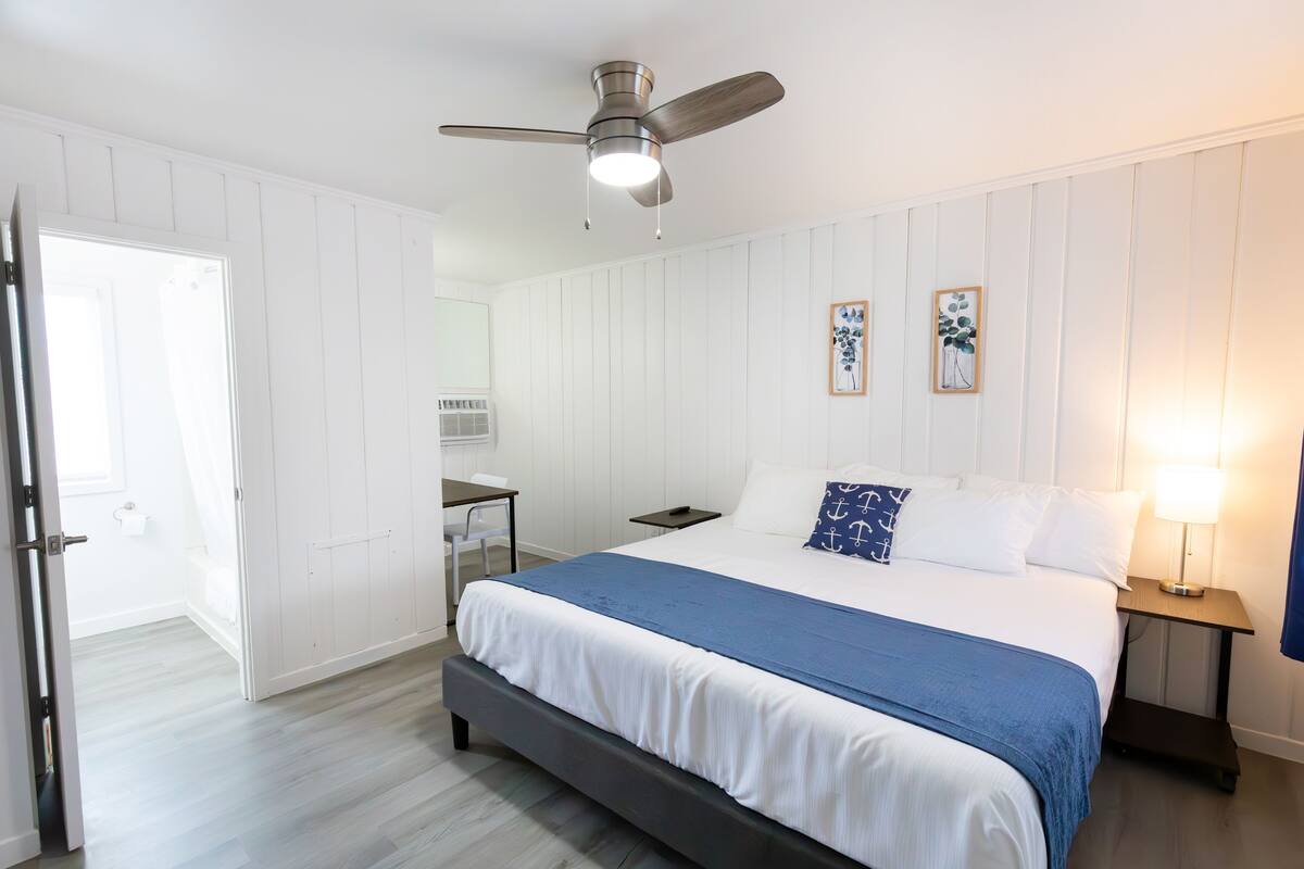 A bedroom with a bed, a ceiling fan, a bedside table with a lamp, and a small table with a chair.