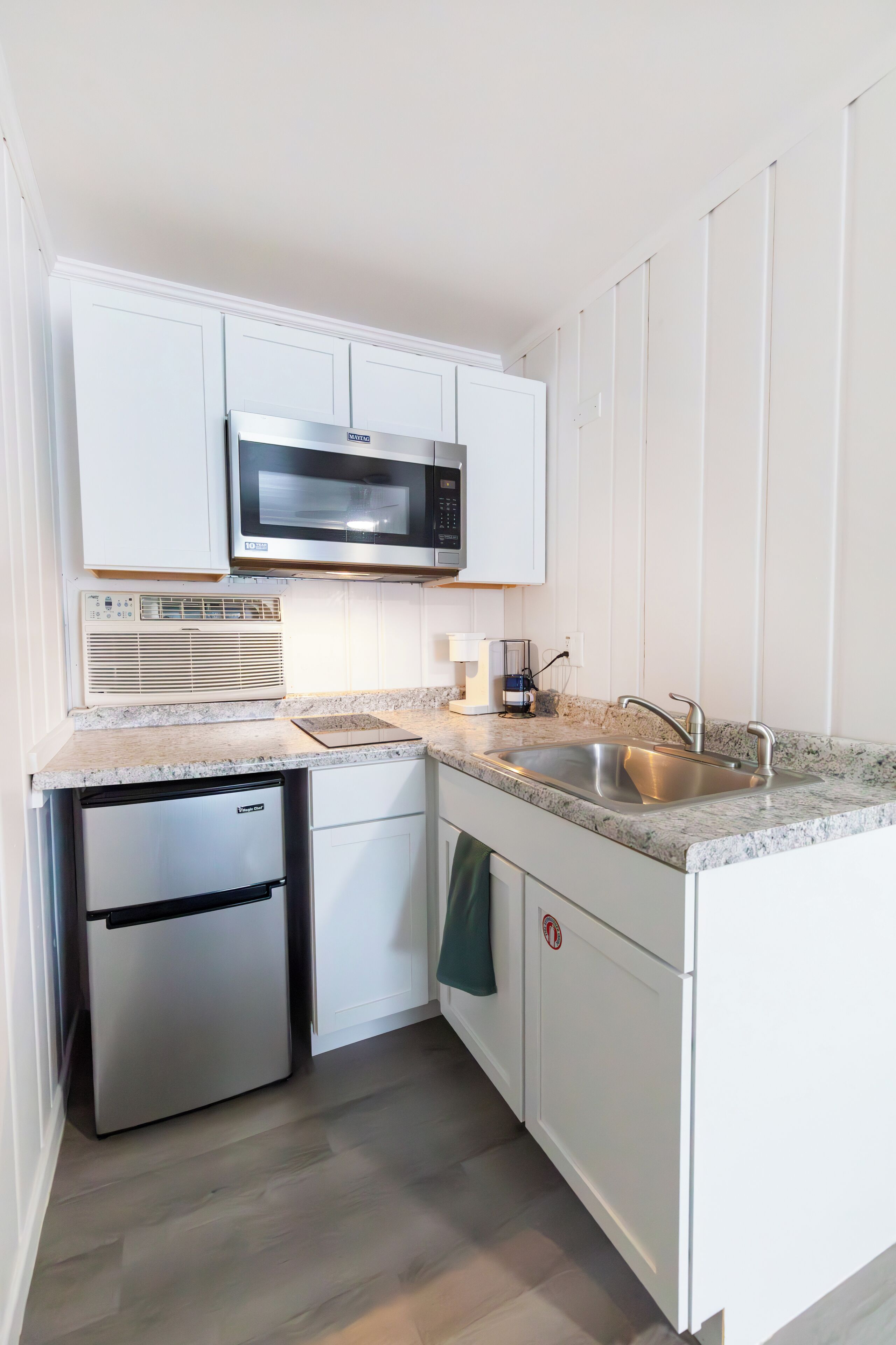 Una cocina compacta con muebles blancos, un microondas de acero inoxidable, horno empotrado, un lavamanos y un lavavajillas pequeño.
