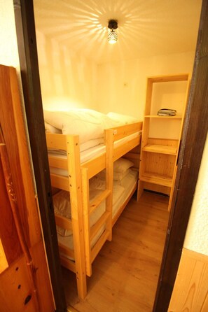 Ein kleiner, schmaler Raum mit einem Etagenbett, einem Holzregal und einer Deckenleuchte.