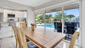 Una cocina moderna con armarios blancos y electrodomésticos de acero inoxidable, un comedor con mesa y sillas de madera, y vista a una piscina a través de amplios ventanales correderos.