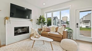 A modern living room with a fireplace, a large TV, a brown sofa, and a large window. - Promenade | 3BR | BBQ, Fire Pit | Near the Beach!! (Sylvan Lake)