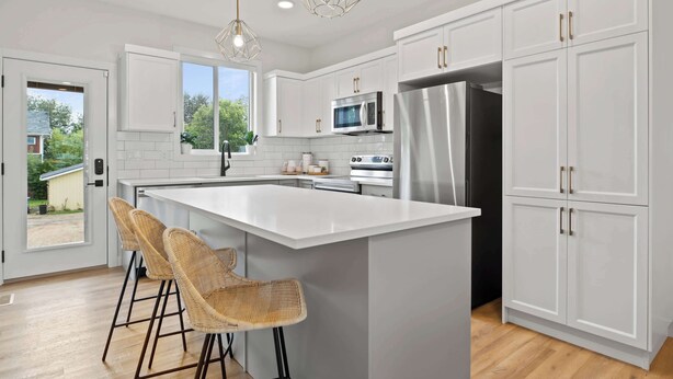 A modern kitchen with white cabinets, a large island, stainless steel appliances, wicker bar stools, and pendant lighting. - Promenade | 3BR | BBQ, Fire Pit | Near the Beach!! (Sylvan Lake)