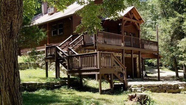 Une cabane en bois dotée d’un balcon, entourée d’arbres et d’un mur de pierres.