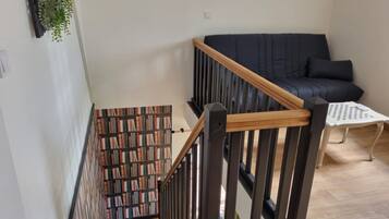 Un escalier avec une rampe en bois, une bibliothèque remplie de livres, un canapé sombre et une petite table sur laquelle est posé un jeu d’échecs.
