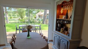 A dining area with a table, chairs, and a vase of flowers. There is a glass door leading to a garden. A cabinet with wine glasses and bottles is visible. - Near Châteaux & zoo de beauval: Gîte de sologne (Pruniers-en-Sologne)