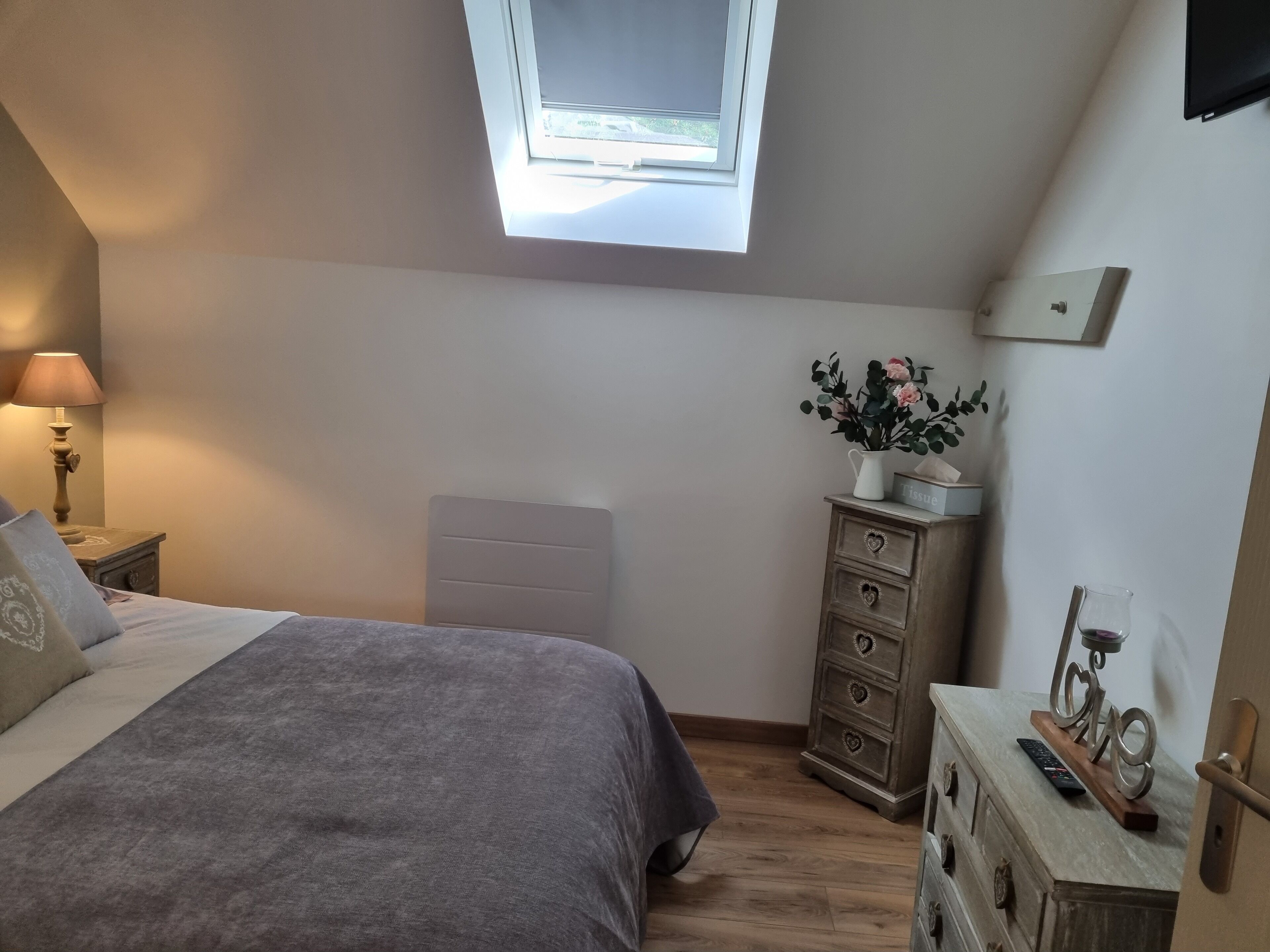 Un dormitorio con cama, cómoda, mesita de noche, lámpara, ventana y una pequeña mesa con un jarrón y un control remoto.