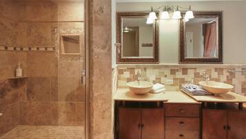 Un baño con ducha envuelta en vidrio, dos lavamanos con cuencos de madera y un tocador con espejo e iluminación superior.