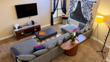 A living room with a flat-screen TV, a wooden entertainment unit, a grey sofa, a coffee table, and a floor lamp.