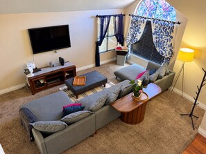A living room with a flat-screen TV, a wooden entertainment unit, a grey sofa, a coffee table, and a floor lamp.