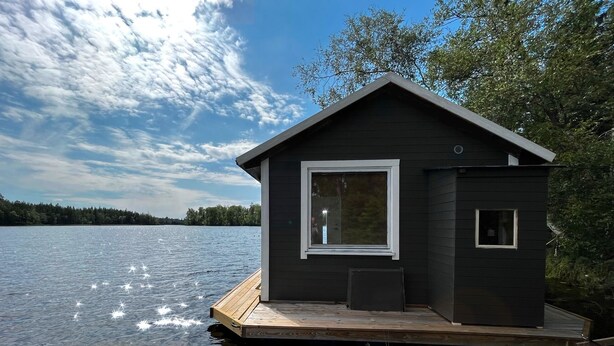 A dark wooden cabin on stilts by a lake with a clear sky. - Cozy Tiny Houseboat in the Nature (Hjulvhult)