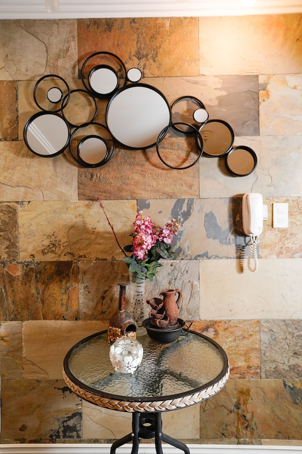 Una mesa de vidrio redonda con un florero decorativo y una pequeña maceta encima, frente a una pared de piedra con un espejo y un teléfono.