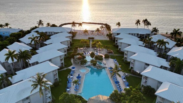 A resort with multiple white buildings, a central pool area with lounge chairs, and a view of the ocean at sunset. - Sunset Cove 2BR/2BA • Ocean Views • Pool/ Swim-Up Bar • Hot Tub • Walk to Beach! (George Town)