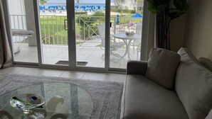 A living room with a glass coffee table, a sofa, and a view of a beach with umbrellas and palm trees. - Sunset Cove 2BR/2BA • Ocean Views • Pool/ Swim-Up Bar • Hot Tub • Walk to Beach! (George Town)