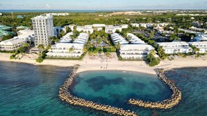 A resort with multiple white buildings, a beach, and a clear blue sea. - Sunset Cove 2BR/2BA • Ocean Views • Pool/ Swim-Up Bar • Hot Tub • Walk to Beach! (George Town)