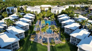 A resort with multiple white-roofed buildings, a central swimming pool, and palm trees. - Sunset Cove 2BR/2BA • Ocean Views • Pool/ Swim-Up Bar • Hot Tub • Walk to Beach! (George Town)